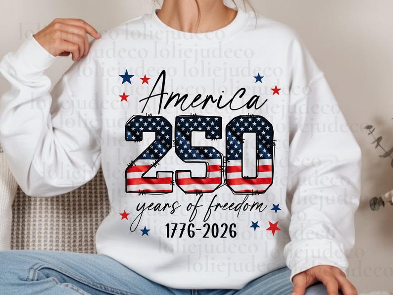 May include: A white long-sleeve sweatshirt featuring a graphic with the text America 250 years of freedom 1776-2026. The number 250 is filled with a red, white, and blue American flag pattern, surrounded by small red and blue stars.