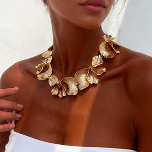 May include: A gold-colored statement necklace with a floral design. The necklace is made of multiple flower-shaped elements. The model is wearing a white tube top.