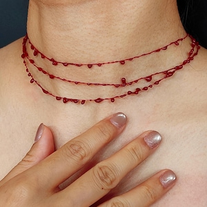 Puede incluir: Un collar rojo de tres hilos con un dise&ntilde;o de goteo de sangre. El collar est&aacute; hecho de hilos rojos finos con gotas en forma de l&aacute;grima espaciadas uniformemente. El collar se lleva alrededor del cuello.