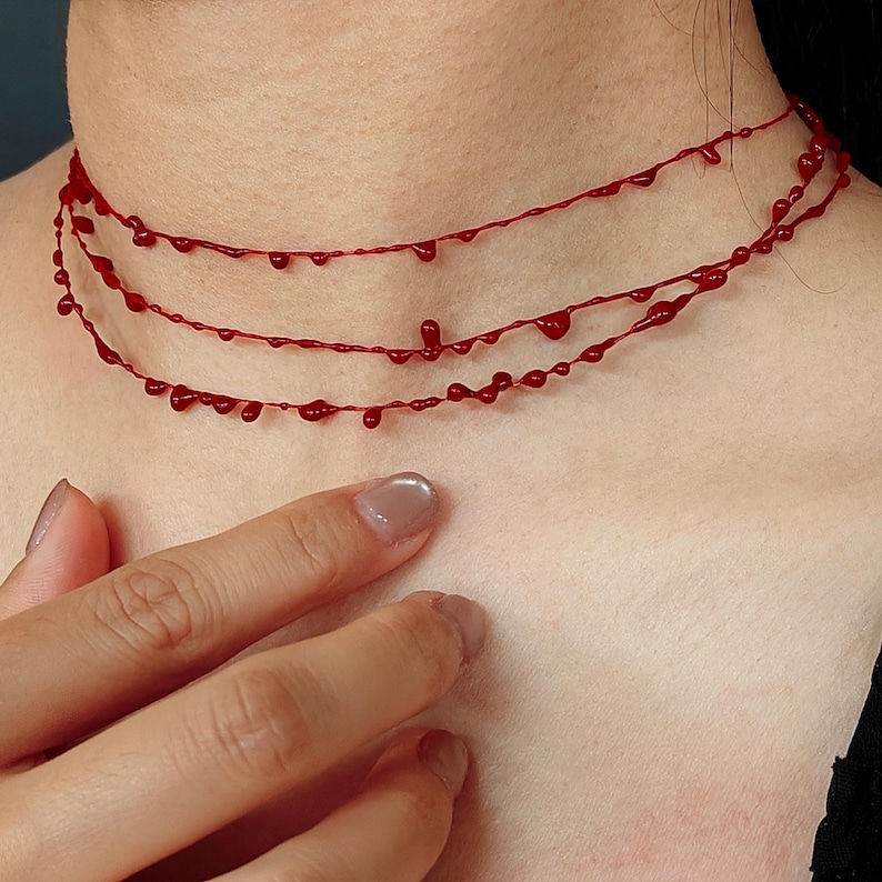 Puede incluir: Un collar rojo de tres hilos con un dise&ntilde;o de goteo de sangre. El collar se lleva alrededor del cuello y presenta una serie de gotas rojas. El collar es una pieza de joyer&iacute;a &uacute;nica.