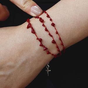 May include: Red beaded bracelet with a heart-shaped design. The bracelet is strung on a red cord and features a small silver cross charm. The bracelet is shown on a wrist against a dark background.