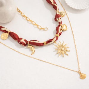 Op de afbeelding: Een meerlagige ketting met een bordeauxrode en cr&egrave;mekleurige stoffen streng en gouden bedels. De bedels zijn onder andere een zon, maan, hart en munt. Een fijne gouden ketting met een schelpenhanger maakt het ontwerp compleet.