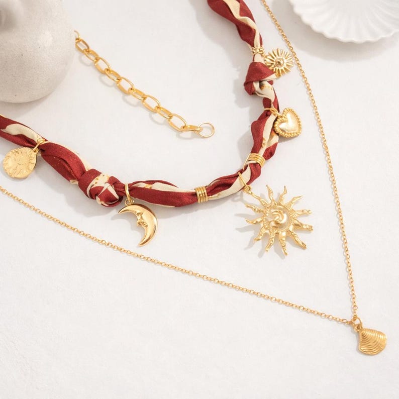 Op de afbeelding: Een meerlagige ketting met een bordeauxrode en cr&egrave;mekleurige stoffen streng en gouden bedels. De bedels zijn onder andere een zon, maan, hart en munt. Een fijne gouden ketting met een schelpenhanger maakt het ontwerp compleet.