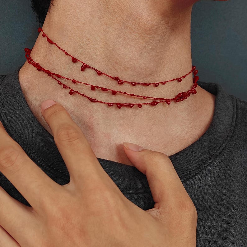 Puede incluir: Un collar gargantilla rojo con un dise&ntilde;o de goteo de sangre. El collar presenta tres hilos de material rojo, cada uno adornado con gotas en forma de l&aacute;grima espaciadas uniformemente. El collar se lleva alrededor del cuello.