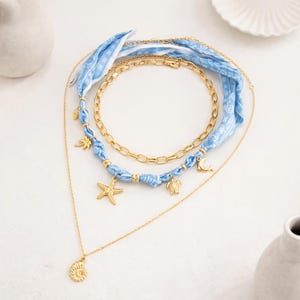 Op de afbeelding: Een gelaagde ketting set met gouden kettingen en bedels. De set bevat een gouden ketting met een schelpenhanger, een gouden ketting en een ketting met blauwe stof en gouden bedels, waaronder een zeester en een schildpad.