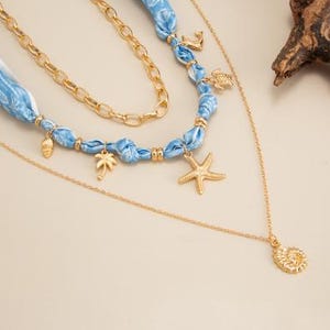 Op de afbeelding: Een meerlagige ketting met gouden kettingen en blauwe stoffen accenten. De ketting heeft bedels, waaronder een zeester, palmboom, zeeschelp en anker. Het ontwerp roept een strand- of nautisch thema op.