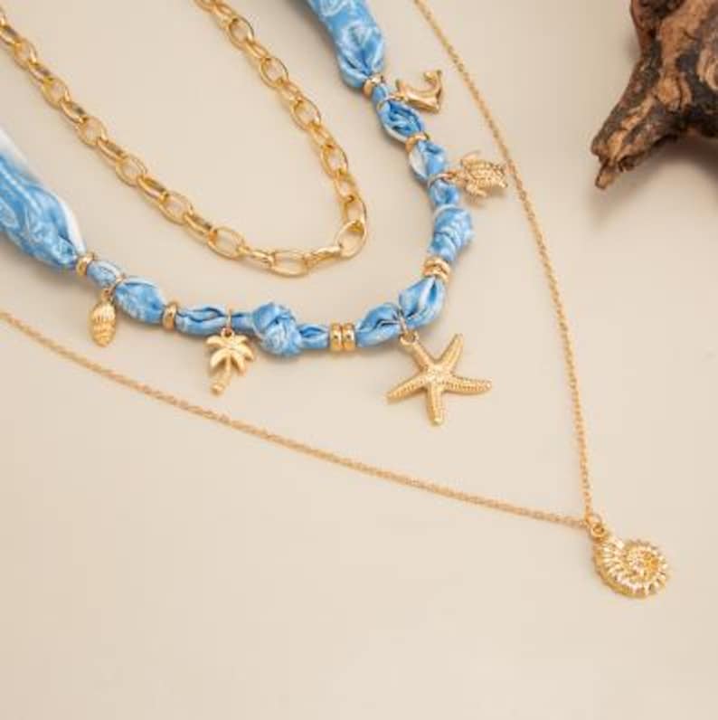 Op de afbeelding: Een meerlagige ketting met gouden kettingen en blauwe stoffen accenten. De ketting heeft bedels, waaronder een zeester, palmboom, zeeschelp en anker. Het ontwerp roept een strand- of nautisch thema op.