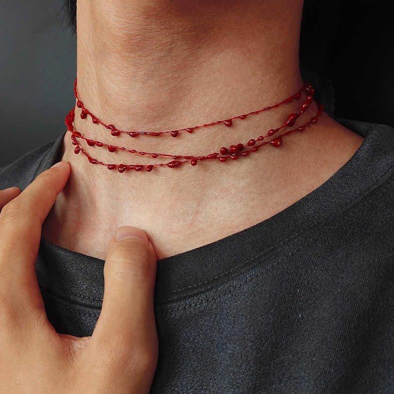 Puede incluir: Un collar rojo con tres hilos, cada uno adornado con peque&ntilde;as cuentas en forma de gota. El collar se lleva alrededor del cuello, creando un efecto de capas. Las cuentas son de un rojo intenso y vibrante.