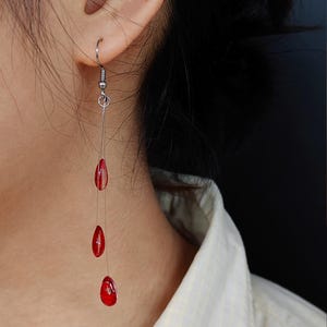 Puede incluir: Pendientes colgantes con tres cuentas rojas en forma de lágrima suspendidas de un gancho plateado. Las cuentas son de un rojo vibrante y parecen ser de vidrio. El fondo es neutro.
