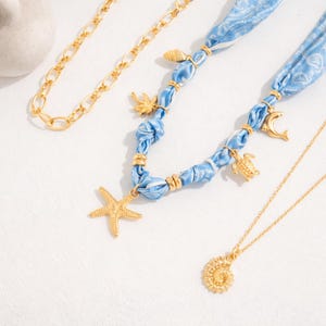 Op de afbeelding: Een verzameling gouden kettingen en een blauwe stoffen ketting met gouden bedels. De gouden kettingen omvatten een schakelketting en een delicate hangerketting met een spiraalvormige schelp. De blauwe ketting heeft een zeester, schildpad en andere bedels.