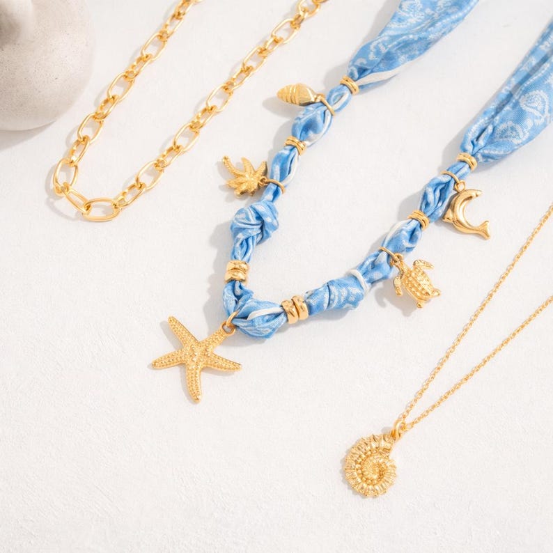 Op de afbeelding: Een verzameling gouden kettingen en een blauwe stoffen ketting met gouden bedels. De gouden kettingen omvatten een schakelketting en een delicate hangerketting met een spiraalvormige schelp. De blauwe ketting heeft een zeester, schildpad en andere bedels.