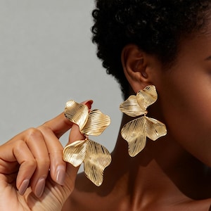 May include: Gold-toned statement earrings featuring a layered, leaf-inspired design. The earrings have a textured surface with visible lines. The earrings are a bold, eye-catching accessory, shown against a neutral background.