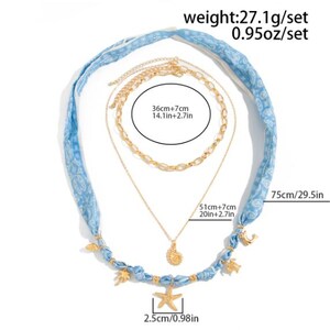 Op de afbeelding: Een gelaagde ketting set met een blauwe stoffen band en goudkleurige kettingen. De set bevat een zeester hanger (2,5cm), een schelp hanger en een ketting van 51cm+7cm lang. De stoffen band is 75cm lang. Het setje weegt 27,1g.