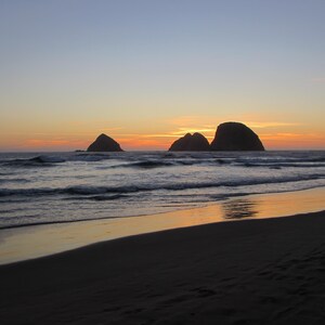 May include: A scenic beach at sunset. The sky transitions from blue to orange and yellow, reflecting on the wet sand. Silhouetted rock formations are in the ocean. Gentle waves roll towards the shore.