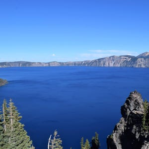 May include: A scenic landscape featuring a large, deep blue lake surrounded by cliffs and evergreen trees. The sky is a clear, bright blue. The image captures a natural environment, possibly a national park or wilderness area.