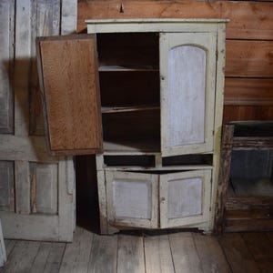 May include: An antique, off-white cabinet with two doors and a wooden interior. One door is open, revealing shelves. The cabinet has a distressed finish, suggesting age and use. The cabinet is in a room with wooden walls and a wooden floor.