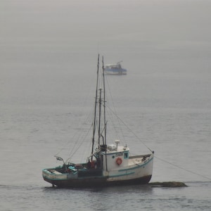 May include: A small, weathered fishing boat with a white hull and blue trim floats on calm water. The boat has a tall mast and rigging, with a second boat visible in the distance. The scene is overcast.