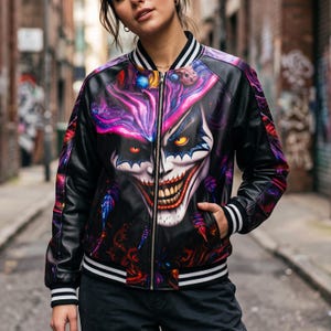 Könnte beinhalten: Eine schwarze Bomberjacke mit einem lebendigen, farbenfrohen Grafikdruck eines finsteren Clown-Gesichts mit leuchtend gelben Augen und einem breiten, zahnigen Grinsen. Die Jacke hat einen Frontreißverschluss, lange Ärmel und schwarz-weiß gestreifte Bündchen.