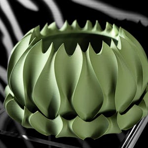 May include: A light green, decorative bowl with a unique petal-like design. The bowl has a textured surface and a scalloped rim. The background is black with a white, abstract pattern. The bowl is displayed on a clear, glass-like surface.