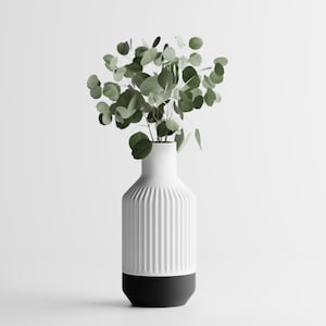 May include: A white ceramic vase with vertical ribbed detailing and a black base, holding a collection of green eucalyptus leaves. The vase has a modern, minimalist design, perfect for home decor.