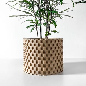May include: A beige, geometric planter with a cylindrical shape, holding a green leafy plant. The planter has a repeating pattern of square cutouts, creating a textured surface. The plant's green leaves contrast with the planter's neutral color.