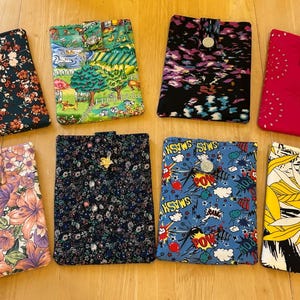 May include: Eight fabric cases with diverse patterns, including florals and landscapes. Each case features a button closure and a unique colour scheme, such as red, blue, and yellow. The cases are displayed on a wooden surface.