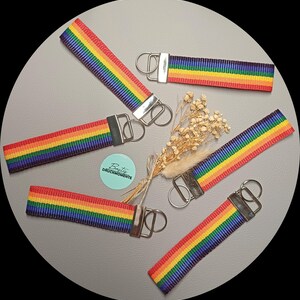 Könnte beinhalten: Fünf Schlüsselanhänger aus Stoff mit horizontalem Regenbogenstreifenmuster in Rot, Orange, Gelb, Grün, Blau und Violett. Jeder Anhänger hat eine silberfarbene Metallklemme und einen Schlüsselring. Ein kleiner türkisfarbener Aufkleber zeigt den Text Bastis DRUCKMOMENTE.