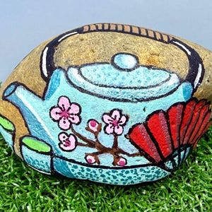 May include: A hand-painted rock featuring a light blue teapot design with pink cherry blossoms and a red fan. The rock has a tan background and black accents. The artwork is set against a backdrop of green artificial grass.
