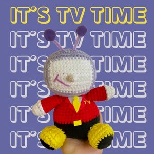 Puede incluir: Muñeco de personaje de televisión tejido a ganchillo, con pantalla blanca, antenas moradas y chaqueta roja. El muñeco tiene pies amarillos y corbata amarilla. El fondo repite las palabras "IT'S TV TIME".