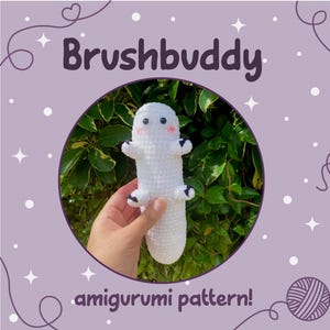 Puede incluir: Una figura amigurumi blanca de ganchillo con detalles negros en los pies y las manos, y mejillas rosadas. La figura se sostiene frente a un fondo de hojas verdes. Se muestra el texto "Brushbuddy" y "amigurumi pattern!"