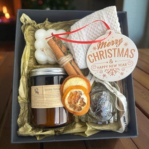 Peut inclure: Un coffret cadeau de Noël avec une bougie, une cannelle, des tranches d'orange séchées, un article tricoté blanc et une étiquette décorative « Merry Christmas & Happy New Year ». Le coffret est garni de papier de soie doré.
