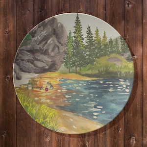 May include: Round watercolor painting depicting a serene lakeside camping scene. The artwork features a campfire, two frogs, and a mug on the shore. The background shows a mountain, evergreen trees, and a calm lake. The color palette includes greens, blues, and browns.