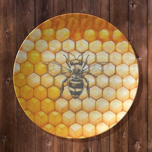 May include: A round decorative plate featuring a detailed illustration of a bee set against a honeycomb pattern. The plate has a yellow and orange color scheme, with a dark gray bee. The plate is likely ceramic or porcelain.