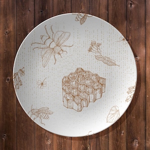 May include: A round, white decorative plate with a brown bee and honeycomb design. The plate features detailed illustrations of bees, honeycomb, and floral elements. The plate is set against a wooden background.