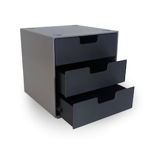 Könnte beinhalten: Graues Aufbewahrungselement mit drei Schubladen und drei schwarzen Schubladen. Die obere Schublade ist geschlossen, während die unteren beiden teilweise geöffnet sind. Das Element ist boxförmig und steht auf einer weißen Oberfläche.