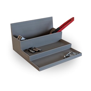 Könnte beinhalten: Ein grauer, gestufter Organizer mit drei Ebenen. Ein rotes Teppichmesser befindet sich im oberen Fach. Der Organizer enthält verschiedene kleine Werkzeuge und Hardware, darunter Schrauben und Bohrer. Das Design ist einfach und funktional.