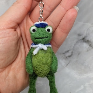 Può includere: Un portachiavi a forma di rana verde in lana infeltrita con un piccolo cappello da marinaio blu e bianco e un colletto da marinaio bianco abbinato con bordo blu e un nodo. La rana ha occhi neri e bianchi ed è fissata a un anello in metallo.