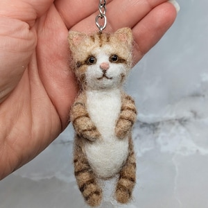 Può includere: Un portachiavi in lana infeltrita a forma di gatto tigrato in piedi con pelliccia a strisce marroni e marrone chiaro, petto e pancia bianchi, piccole orecchie a punta, occhi scuri e naso rosa, attaccato a un anello e catena in metallo argentato.