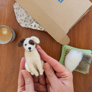 Puede incluir: Figura de perro de fieltro hecha a mano, en colores crema y marrón, sostenida en las manos. El perro tiene una mancha marrón sobre un ojo y orejas marrones. Una caja de manualidades con una pegatina de arcoíris y materiales de manualidades están en el fondo.