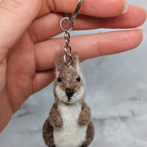 Op de afbeelding: Een naaldgevilte eekhoorn-sleutelhanger met een getextureerd lichaam van bruine en witte wol, kleine zwarte kraaloogjes en een donkere neus. De eekhoorn is met een korte ketting bevestigd aan een zilverkleurige metalen ring als hangend accessoire.
