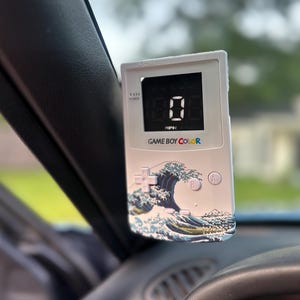 May include: White Game Boy Color device with a digital display showing "0" MPH. The device has a wave design on the front and the words "GAME BOY COLOR" are printed on the front.