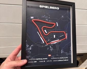 May include: Framed print of the Spielberg race track in Austria. The track is highlighted in red against a dark background. Text at the top reads "SPIELBERG". Additional text provides location, coordinates, and track details. The frame is black.