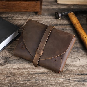 May include: A brown leather wallet with a flap closure and a strap. The wallet has a rustic appearance with visible stitching and a textured surface. The design is simple and classic, suitable for carrying cards and cash.