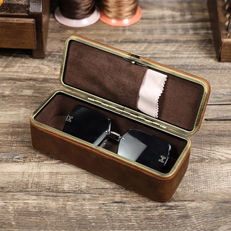 May include: Open brown leather eyeglasses case with a gold-colored frame, lined with brown fabric. Inside, rimless sunglasses with silver-colored arms rest on the fabric. A small white cleaning cloth is tucked inside the case.