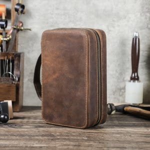 Puede incluir: Un estuche rectangular de cuero marrón con cierre de cremallera y un asa pequeña. El estuche tiene un acabado envejecido y costuras visibles. El fondo incluye herramientas sobre una superficie de madera.