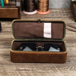 May include: Open brown leather glasses case with a gold-colored frame, lined with brown fabric. Inside, a pair of black rimless sunglasses rests on the case's interior. A white cleaning cloth is tucked inside the case.