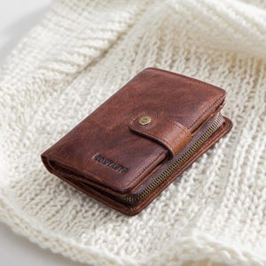 May include: A brown leather wallet with a button clasp and a zip. The wallet is rectangular and has a worn appearance. The word "COWAZIR" is embossed on the front.