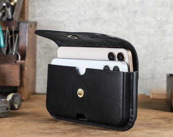 May include: Black leather phone case with a flap and snap closure. The case is open, revealing two smartphones inside. The case has a belt loop on the back and a gold-colored snap closure. The case is sitting on a wooden surface.