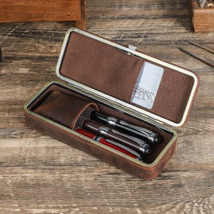 Peut inclure: &Eacute;tui &agrave; stylos en cuir marron ouvert, r&eacute;v&eacute;lant un int&eacute;rieur marron clair. Il contient trois stylos, dont deux avec des d&eacute;tails argent&eacute;s et un avec un corps rouge. Une petite poche contient une carte argent&eacute;e.