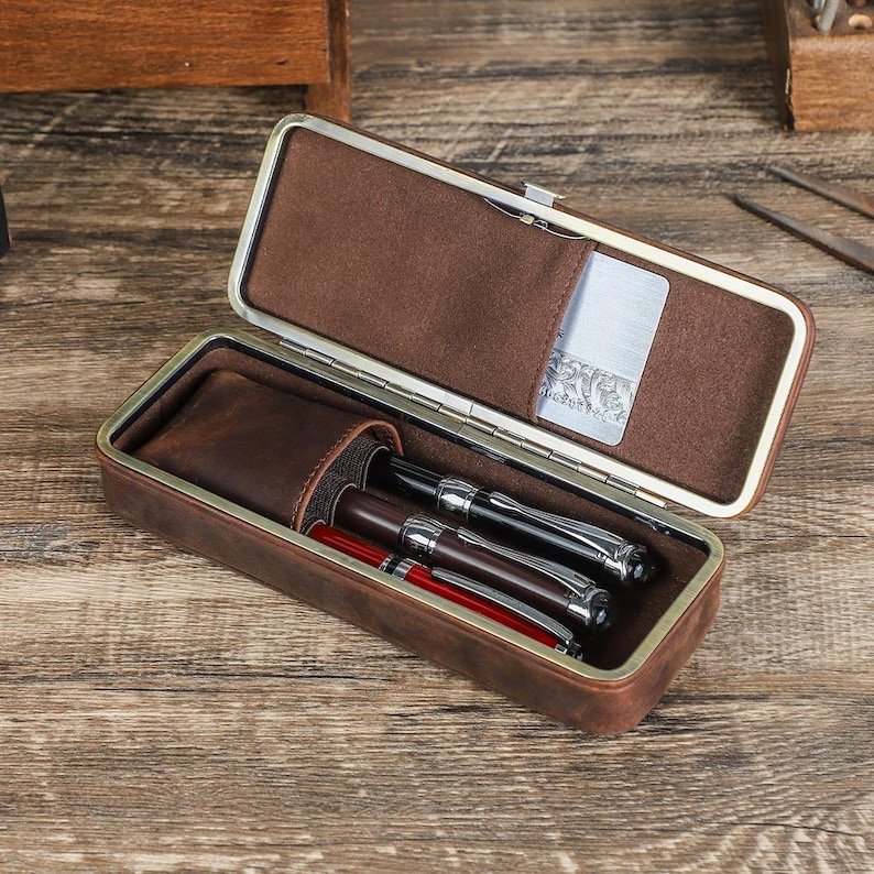 Peut inclure: &Eacute;tui &agrave; stylos en cuir marron ouvert, r&eacute;v&eacute;lant un int&eacute;rieur marron clair. Il contient trois stylos, dont deux avec des d&eacute;tails argent&eacute;s et un avec un corps rouge. Une petite poche contient une carte argent&eacute;e.